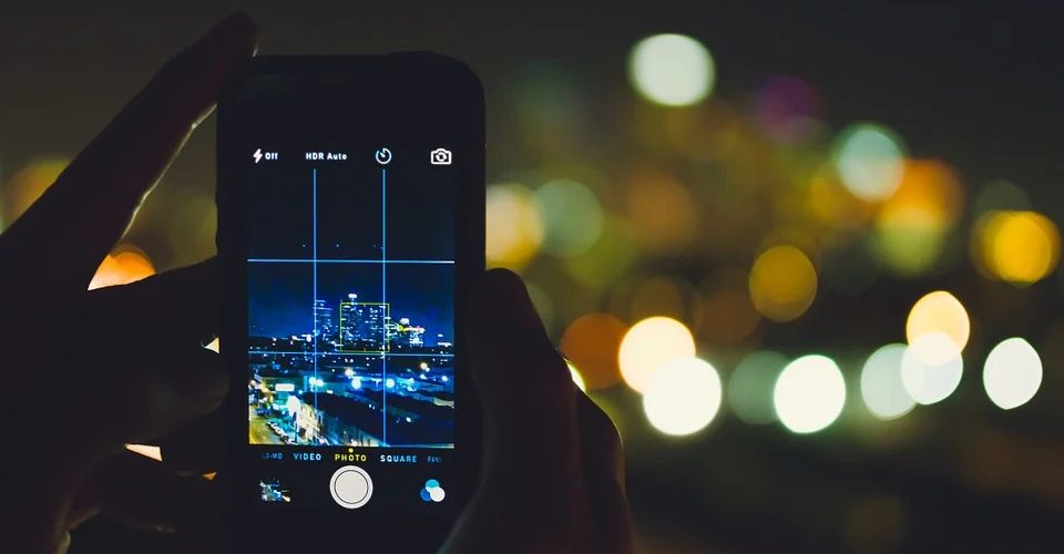 Im Vordergrund wird ein Smartphone mit geöffnetem Kameramodus ins Bild gehalten, durch das Display ist eine Stadt bei Nacht zu sehen
