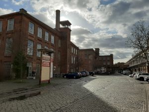 Links Gebäude der Baumwollspinnerei aus Backsteinen, rechts im Bild führt eine gepflasterte, unbefahrene Straße am Gebäude vorbei, an den Seiten parken Autos