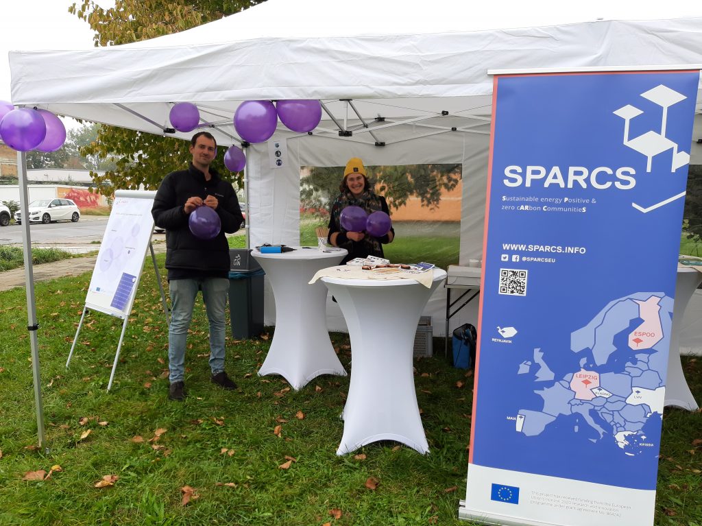 Pavillon, unter dem zwei Stehtische und zwei Personen stehen und in die Kamera lächeln, rechts im Bild ein Roll-Up mit SPARCS-Logo