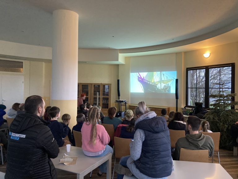 Man sieht einen Raum mit Kindern und Erwachsenen, die auf einer Leinwand einen Film schauen