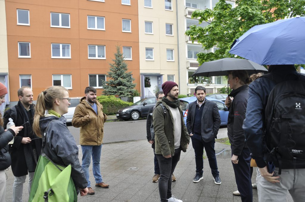 Eine Gruppe besucht das Kreuzstraßenviertel. Man sieht Menschen vor einem sanierten Platten-Gebäude und eine Person, die etwas darüber erzählt.